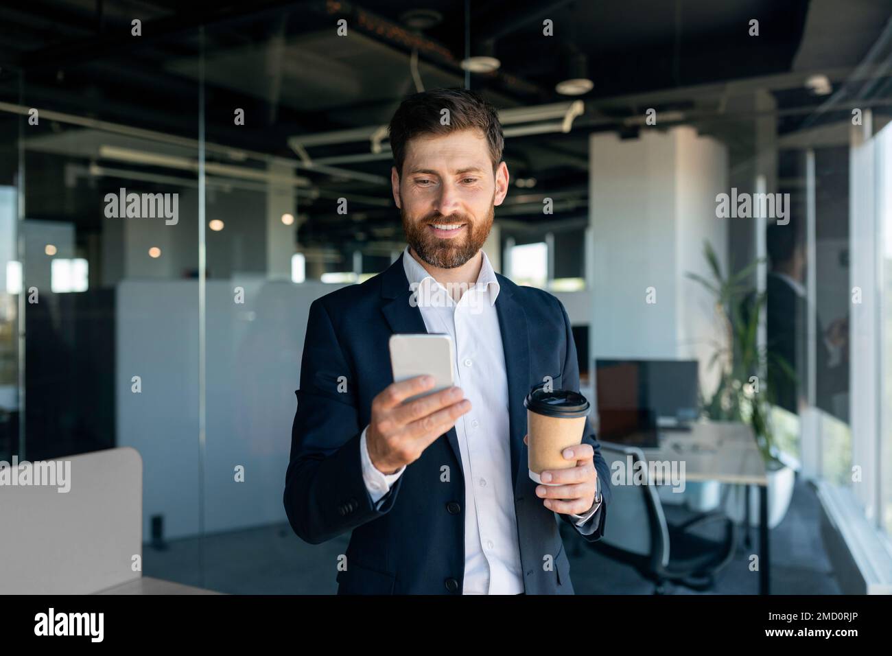 Positive middle aged businessman with beard in suit with cup of coffee using smartphone in office interior, free space Stock Photo