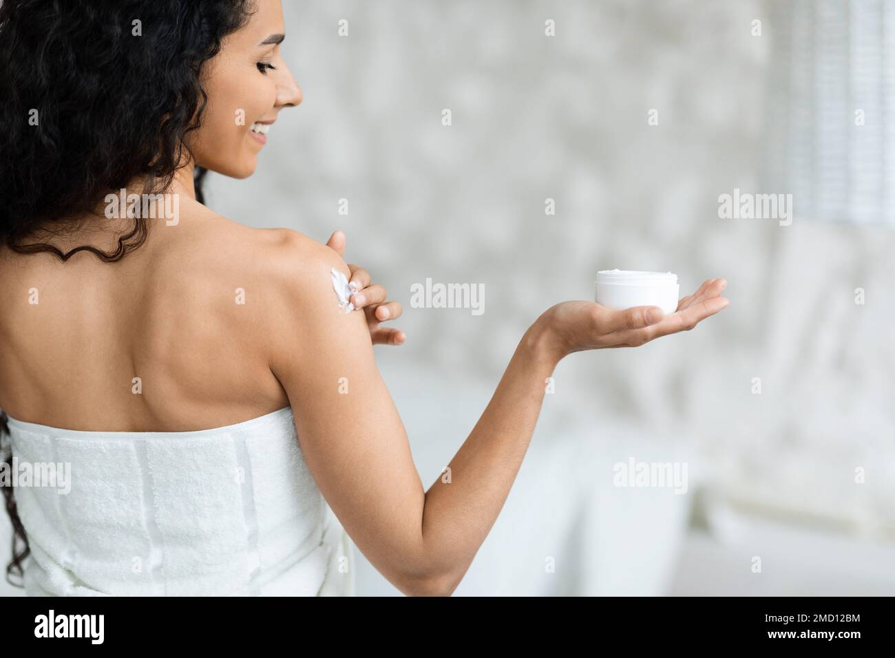 Smiling arabic millennial curly lady in towel holding jar, applies cream on shoulder in bedroom interior, back Stock Photo