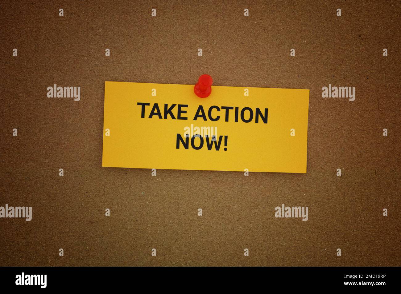 A yellow paper note with the phrase Take Action Now! on it pinned to a cardboard background. Close up. Stock Photo