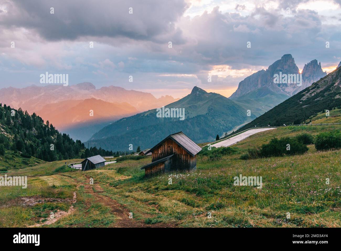 Traditional wooden houses in the Italian Alps in a cinematic landscape with golden light, a mountain road and impressive mountains. Dolomites, Italy. Stock Photo