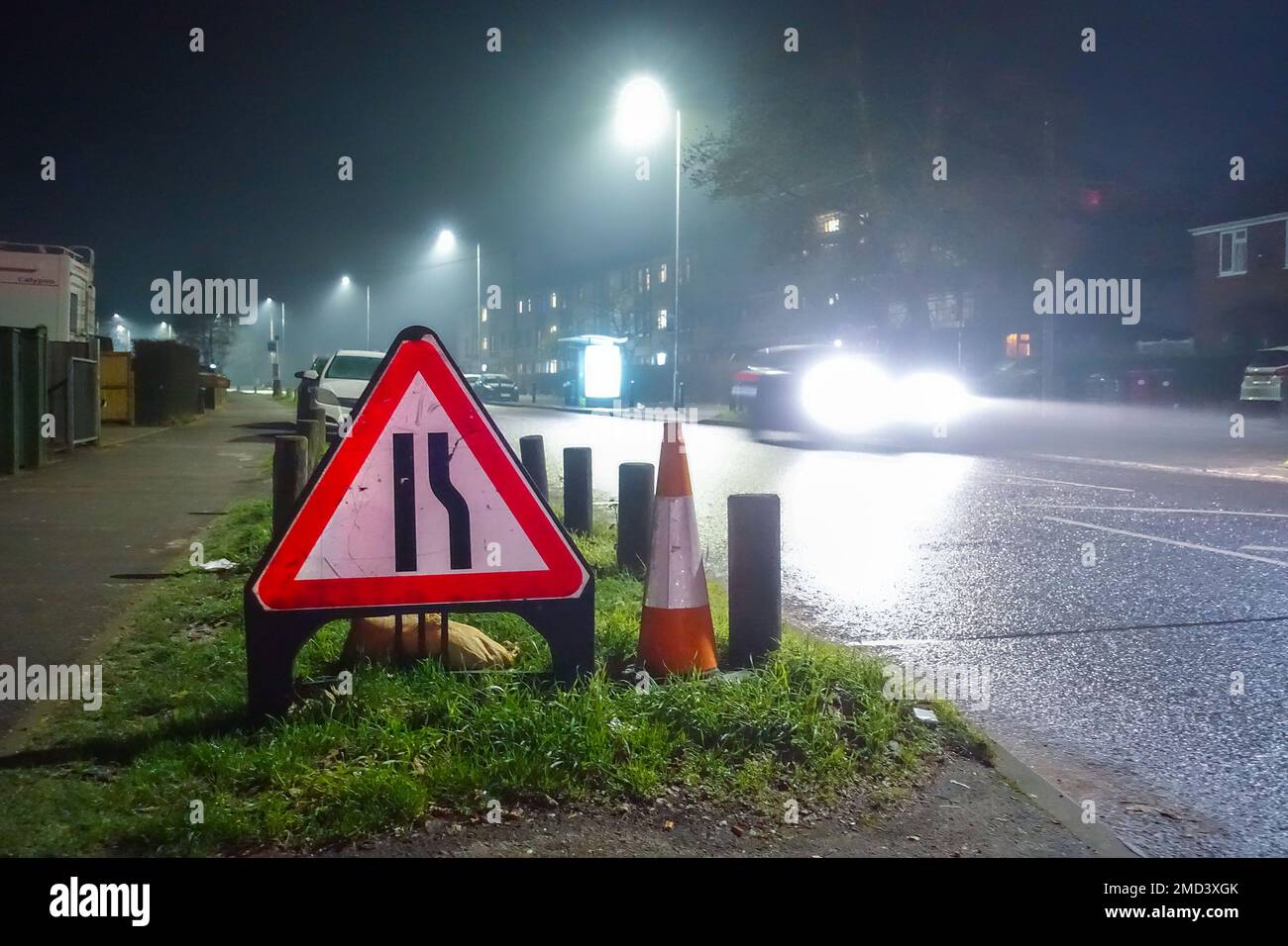 A triangular roadworks sign and traffic cone at the side of a road at night to worn motorists of a narrowed road ahead, Stock Photo