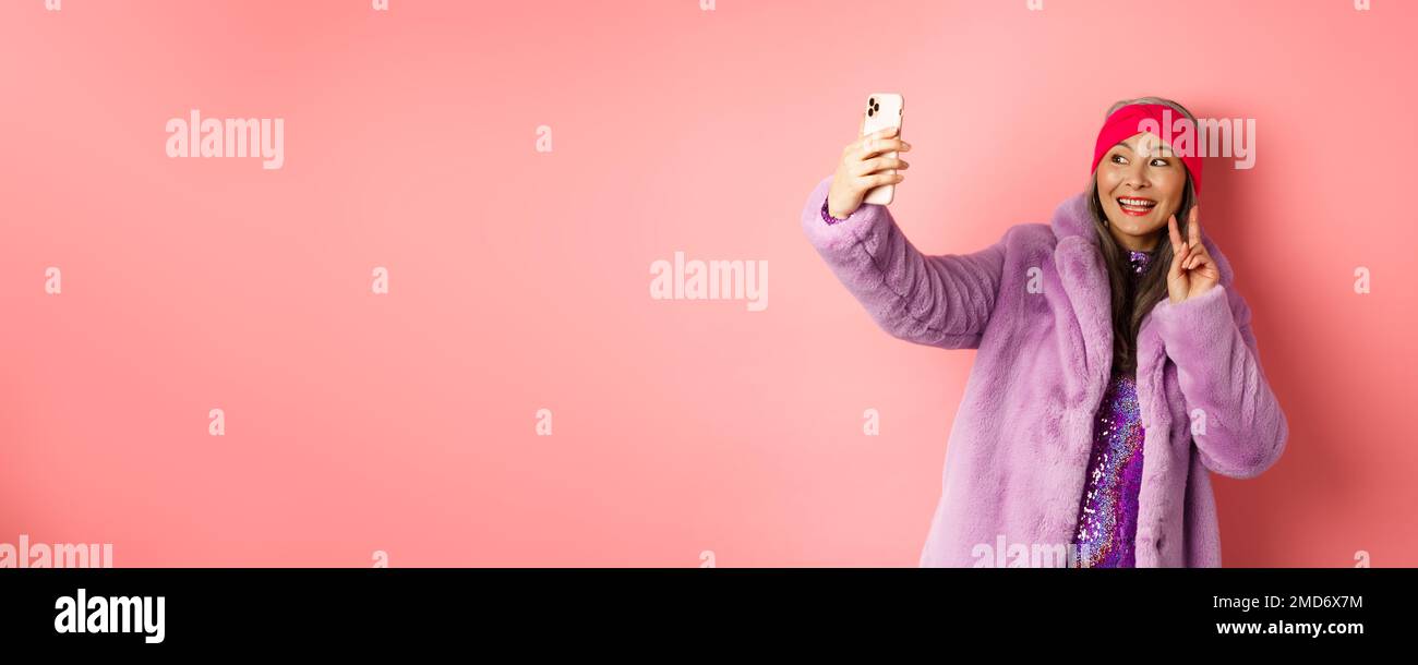 Fashion concept. Cool and cheerful asian senior woman taking selfie with peace sign, wearing trendy faux fur coat and party dress, standing over pink Stock Photo