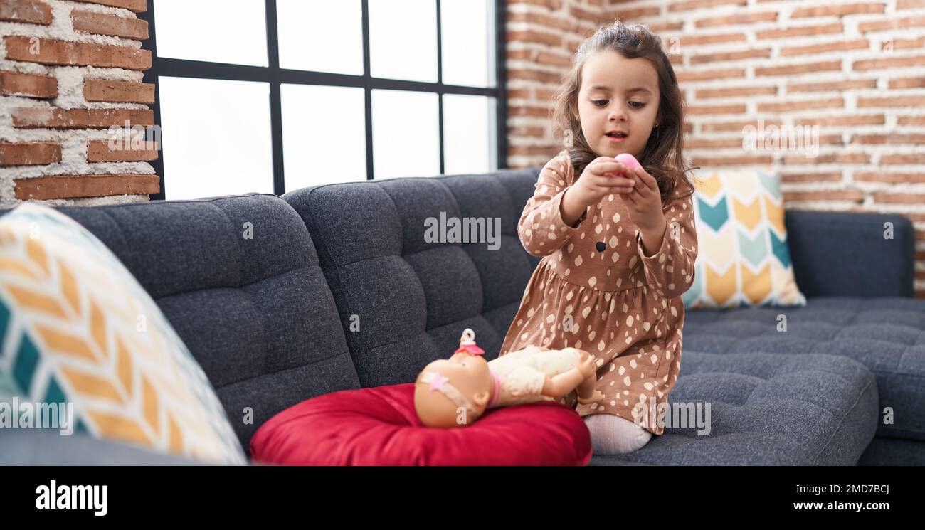 Adorable hispanic girl playing with baby doll sitting on sofa at home ...