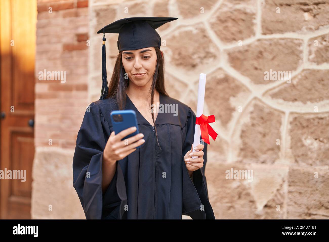 Young hispanic woman wearing graduated uniform holding diploma using ...