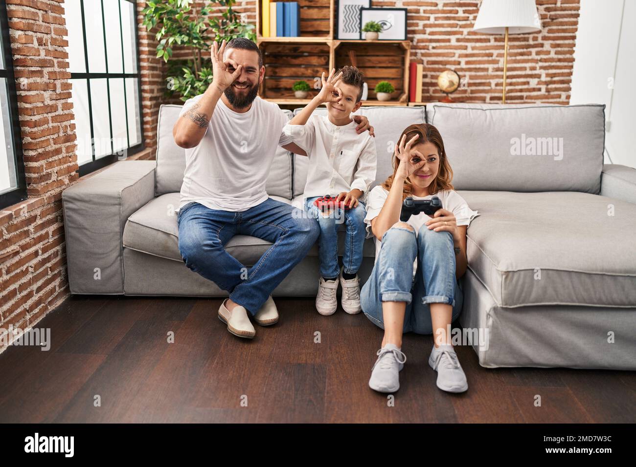 Family of three playing video game sitting on the sofa smiling happy doing ok sign with hand on eye looking through fingers Stock Photo