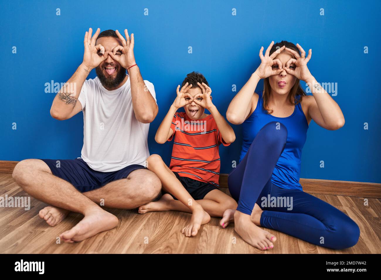 Family of three sitting on the floor at home doing ok gesture like binoculars sticking tongue out, eyes looking through fingers. crazy expression. Stock Photo