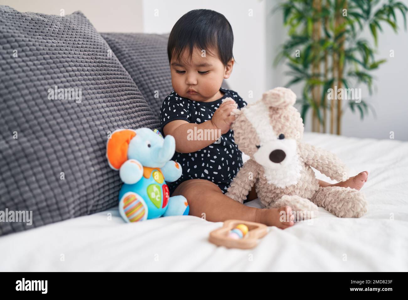 Adorable hispanic baby playing with dolls sitting on bed at bedroom ...