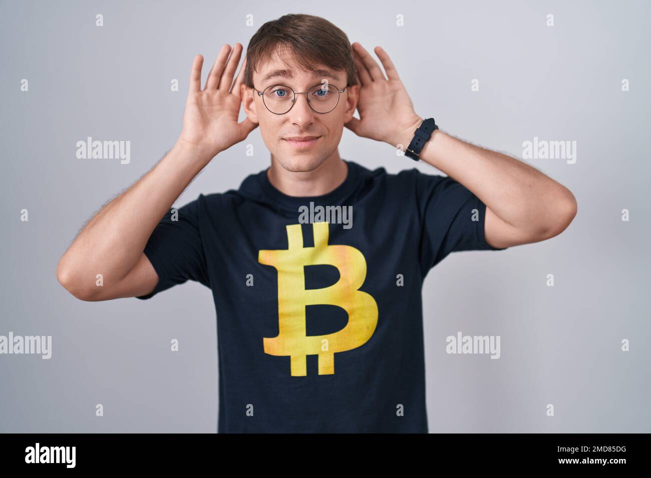 Caucasian blond man wearing bitcoin t shirt trying to hear both hands on  ear gesture, curious for gossip. hearing problem, deaf Stock Photo - Alamy