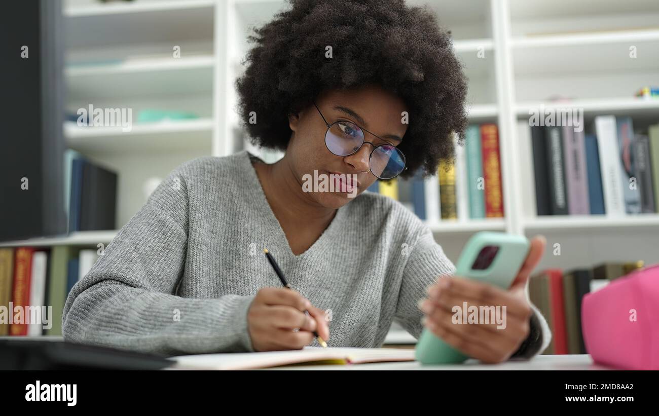 African american woman student using smartphone writing on notebook at ...