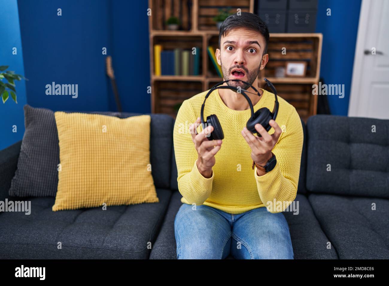 Hispanic man holding broken headphones in shock face, looking skeptical