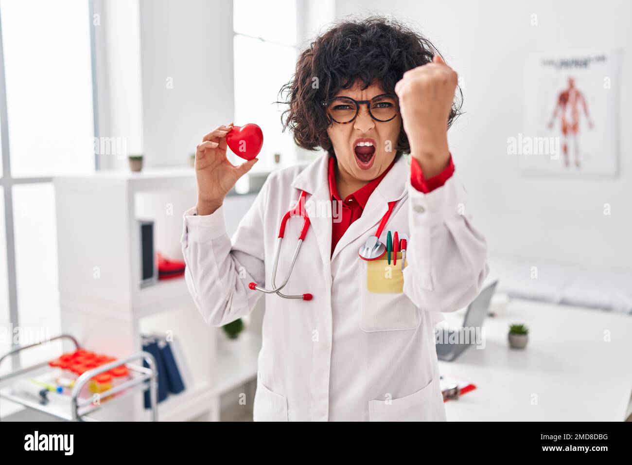 Hispanic doctor woman with curly hair holding heart annoyed and ...