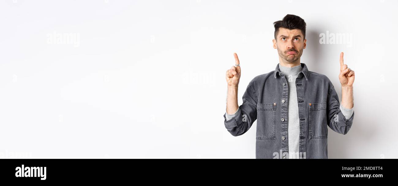 Hesitant Sad frowning guy pointing and looking up, standing hesitant on white ...