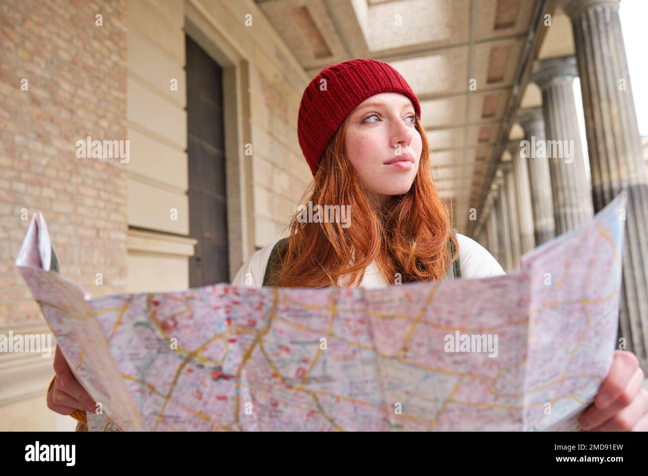 Smiling young redhead woman in red hat, looks at paper map to look for tourist attraction. Tourism and people concept. Girl explores city, tried to Stock Photo