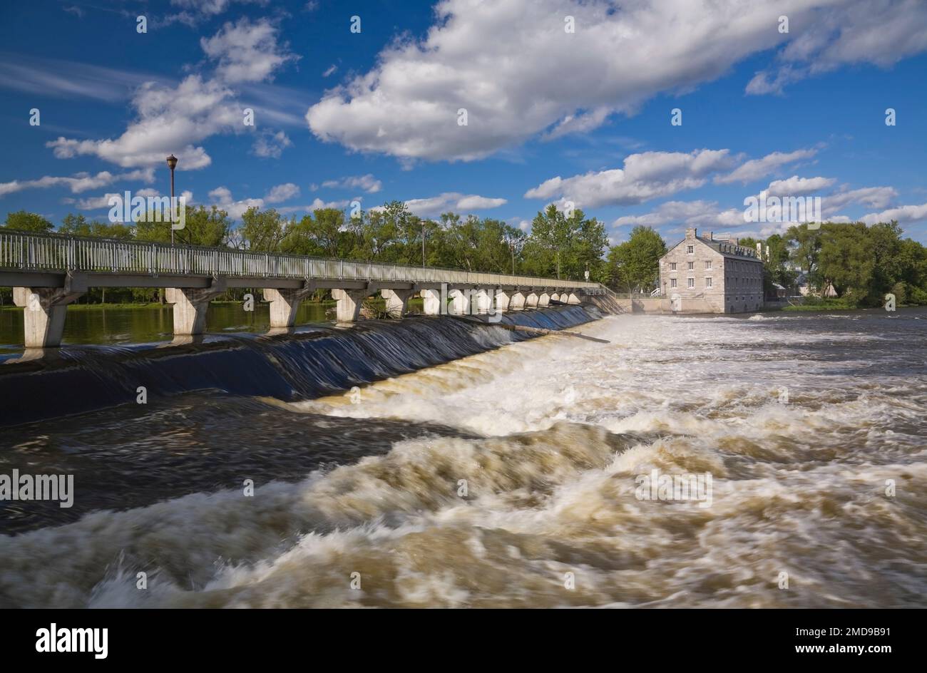 Footbridge and water flow control gate over Des Mille-Iles River and New Mill on Ile des Moulins Historic Site in spring, Old Terrebonne, Quebec. Stock Photo