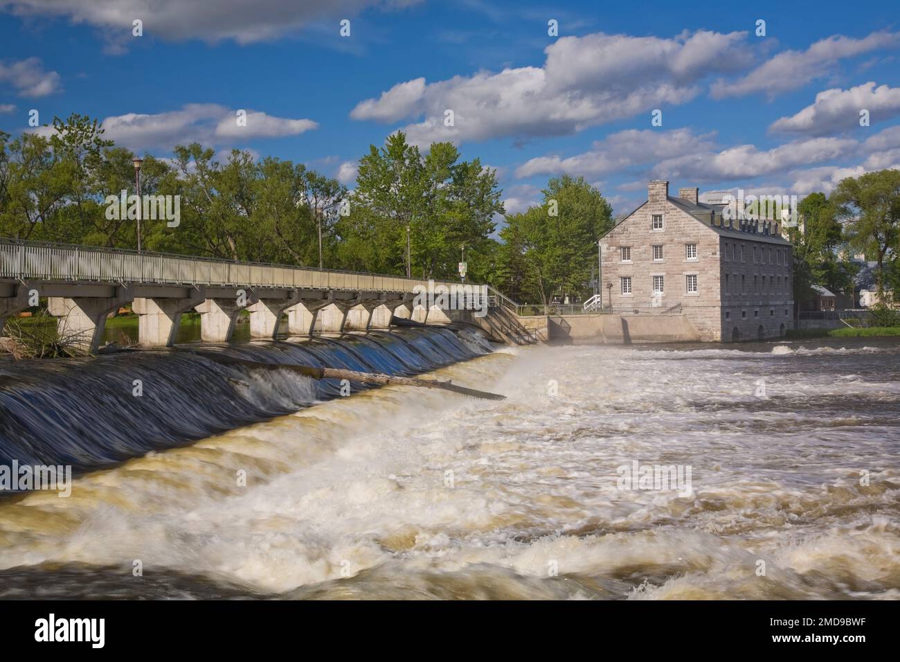 Footbridge and water flow control gate over Des Mille-Iles River and New Mill on Ile des Moulins Historic Site in spring, Old Terrebonne, Quebec. Stock Photo