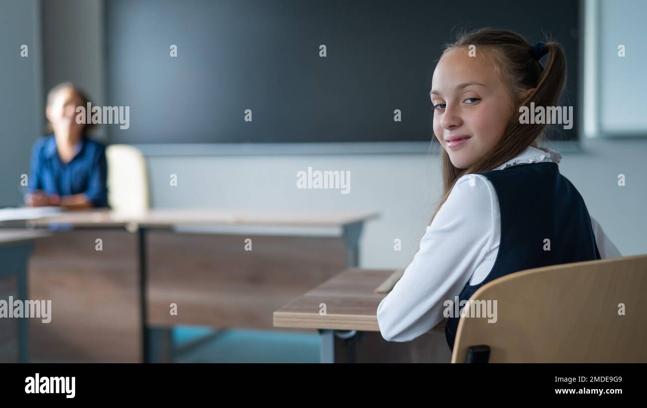 Caucasian girl and her teacher in the classroom. Schoolgirl turns around and looks at the camera.  Stock Photo