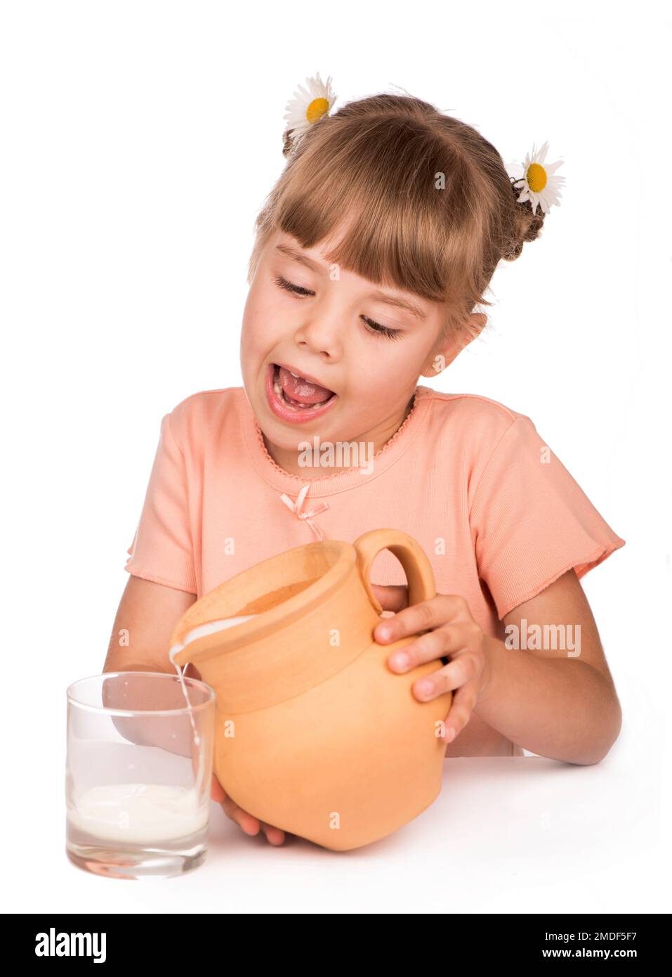 Healthy food. A little girl in an orange T-shirt and flowers in her hair pours fresh milk from a jug on a white background Stock Photo