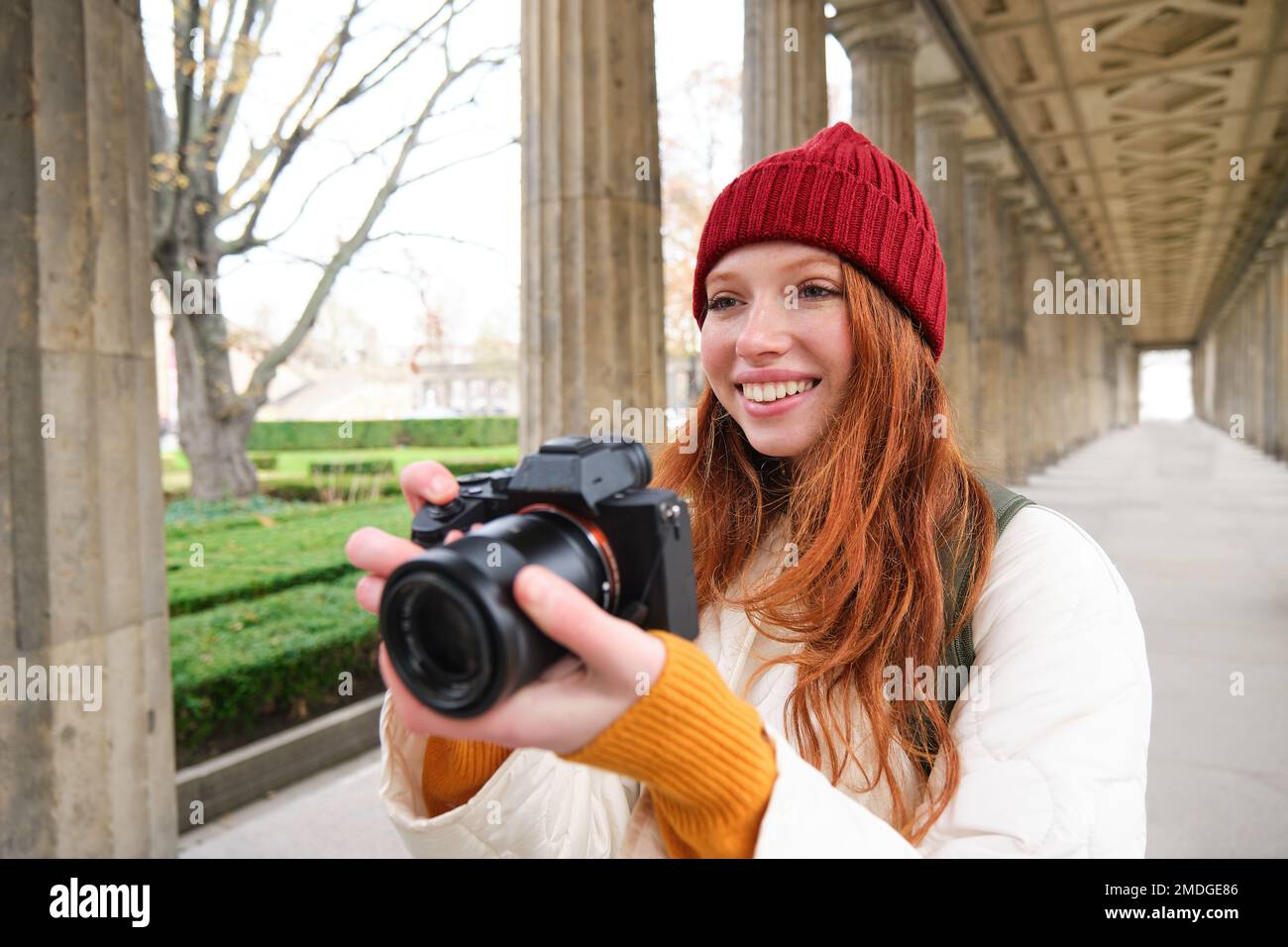Young redhead female photographer, makes lifestyle shooting in city centre, takes photos and smiles, looks for perfect shot, makes picture Stock Photo