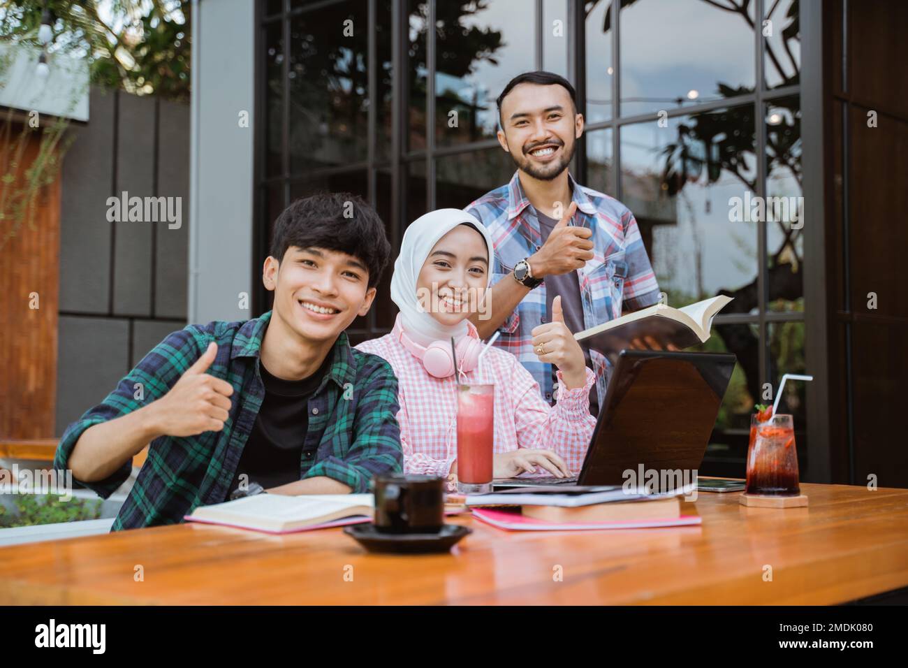 three smiling Asian students with thumbs up while studying together ...