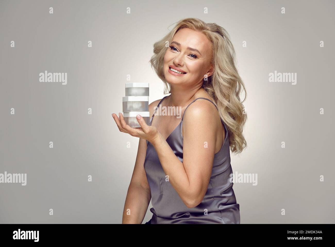 Caucasian mature woman holding jars of moisturizer near her face on a grey isolated. Stock Photo