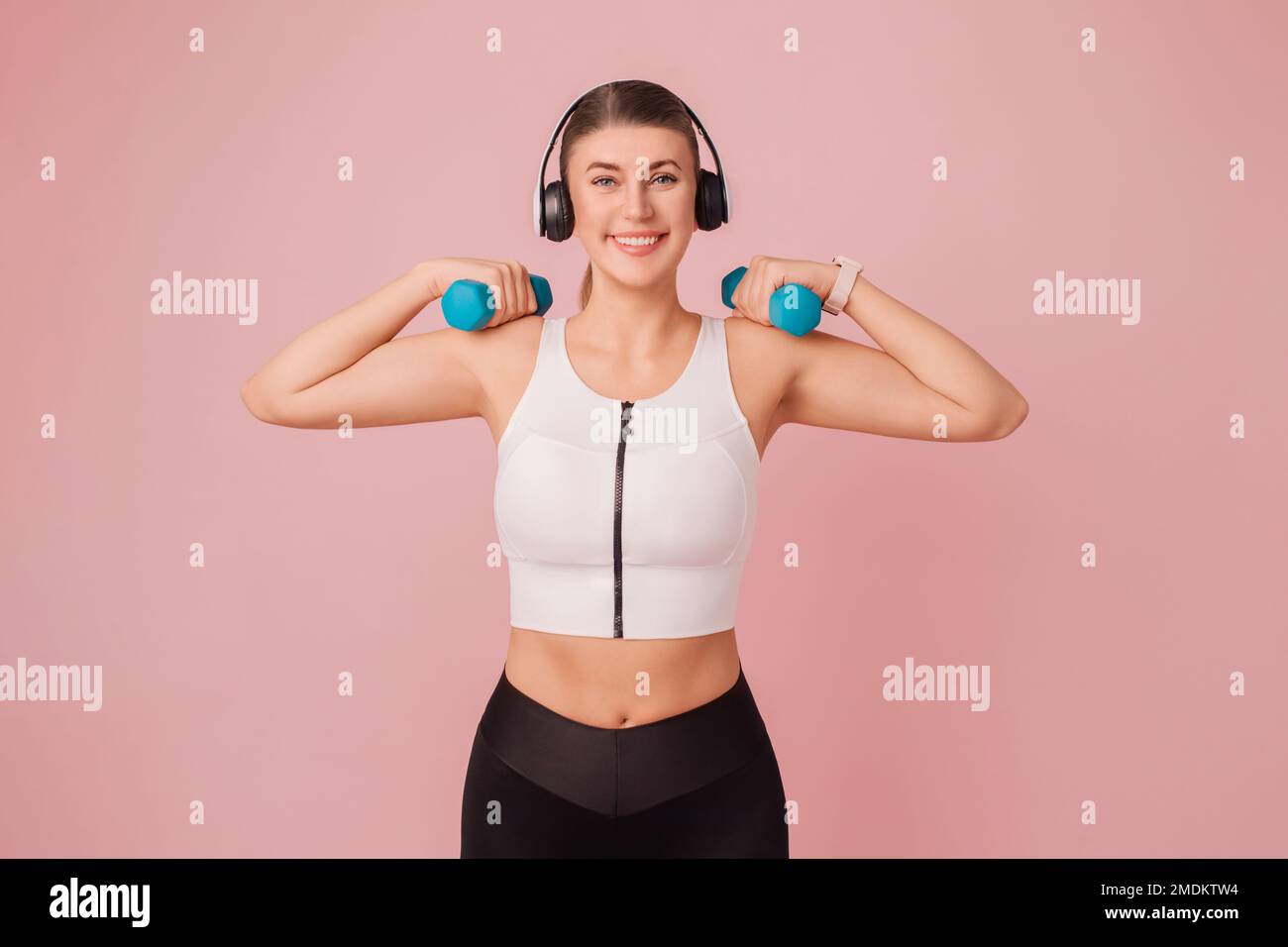 Happy smiling caucasian fitness woman lifting blue dumbbells. Attractive young woman wearing sportswear doing sport exercises over pink background Stock Photo