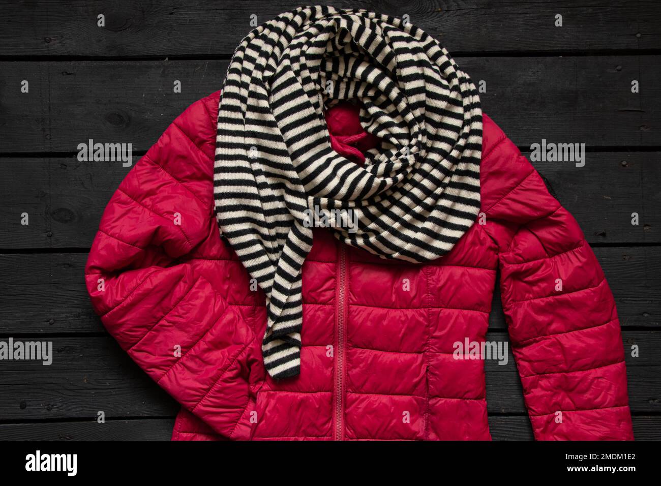 red winter women's warm jacket and striped black and white scarf lie on a black wooden table, winter clothes, jacket Stock Photo