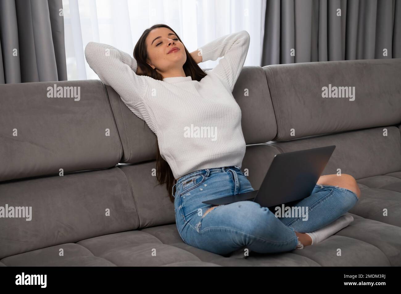 Relaxed young woman lounges on a comfortable sofa, arms crossed behind her head and holding laptop on her knees in a cozy modern living room.  Stock Photo