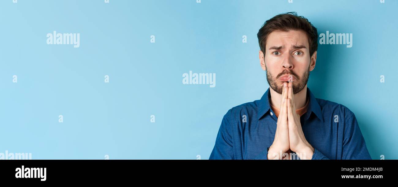 Sad young man begging for help, apoligizing and sobbing miserable, standing on blue background Stock Photo