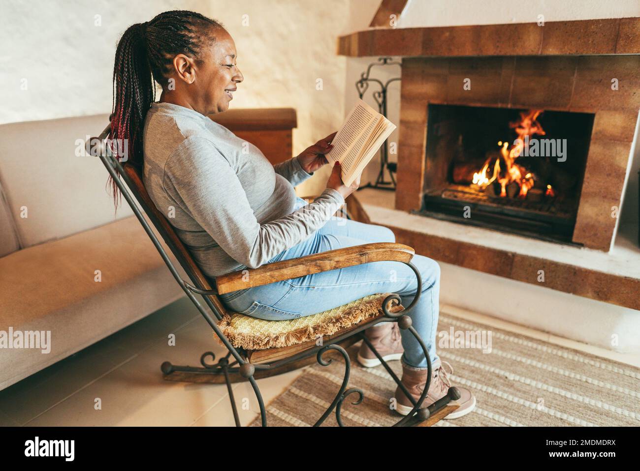 African woman reading book in front of cozy fireplace at home - Winter lifestyle concept - Focus on face Stock Photo