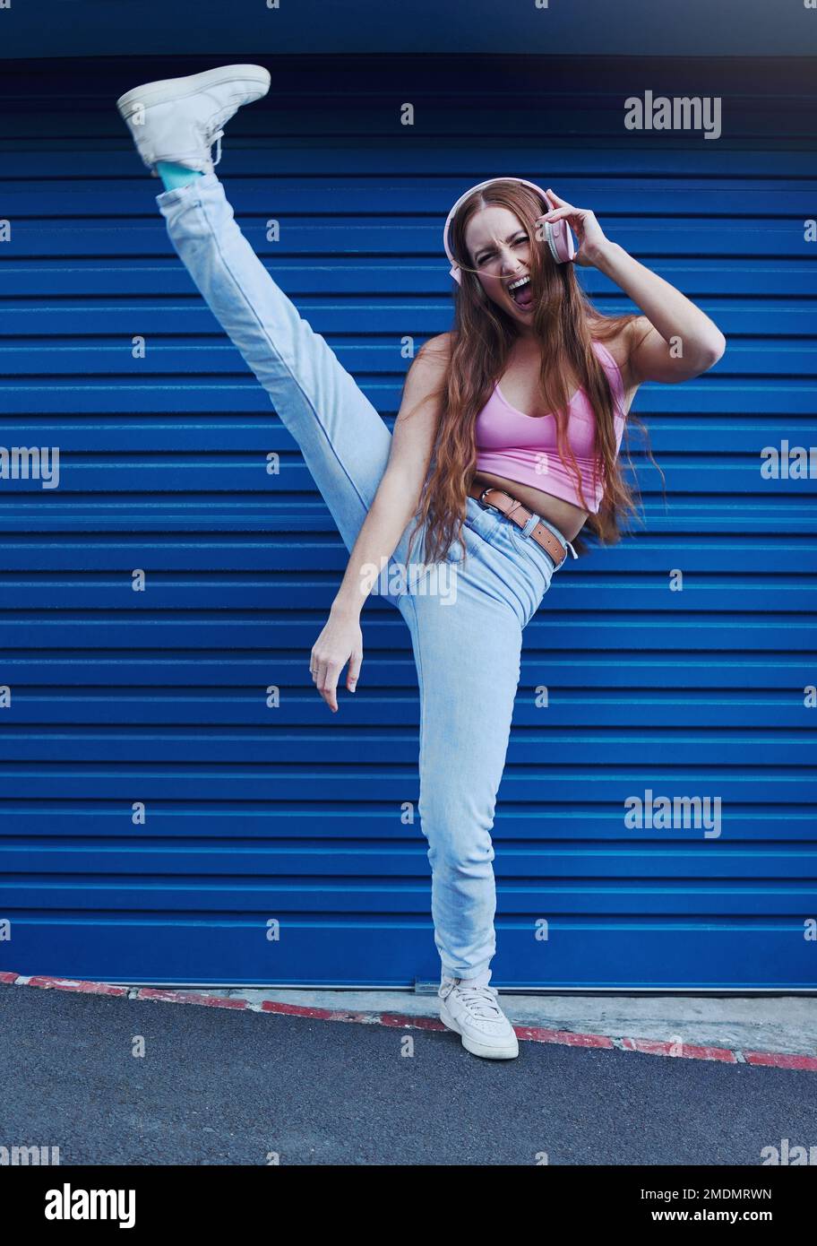 Crazy music, freedom and excited girl on blue background in city listening to track and audio on headphones. Fashion, lifestyle and woman with leg up Stock Photo