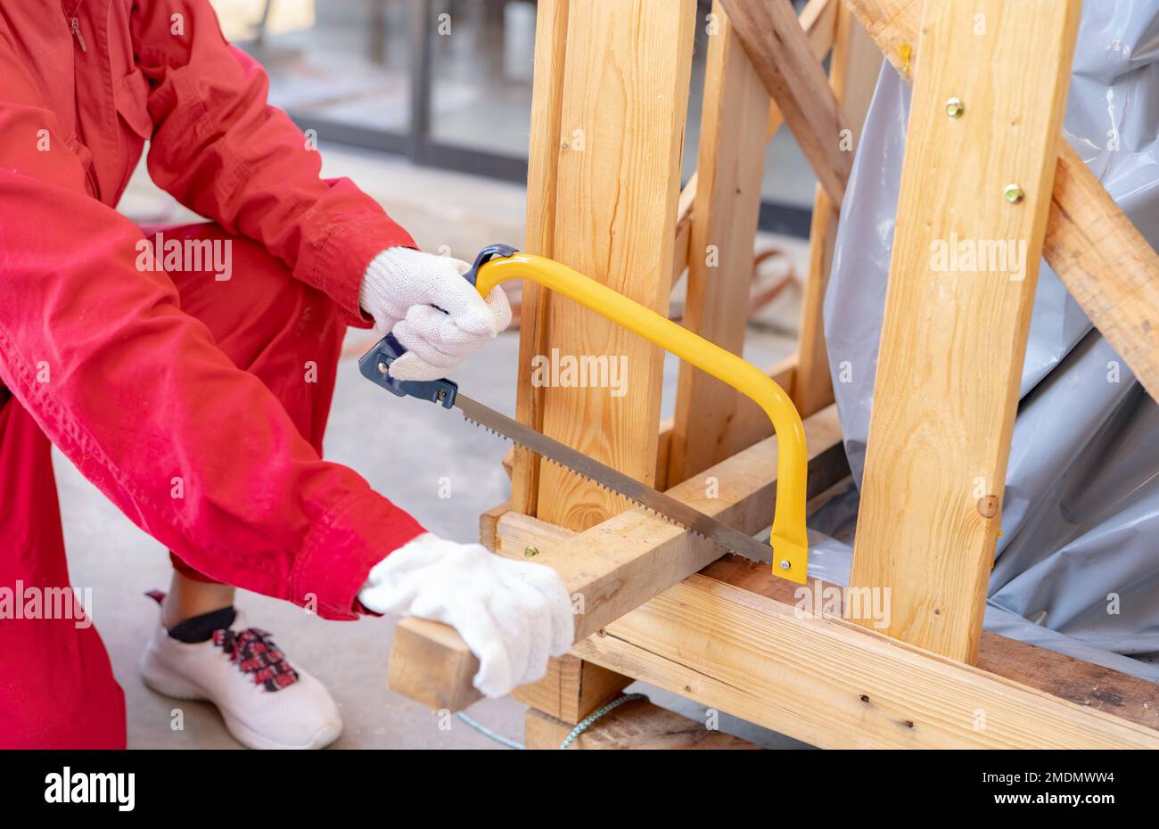 A worker is sawing wood to assemble a crate for moving an industrial machine. A woman in red mechanic coveralls hand holding a bow saw cutting Stock Photo
