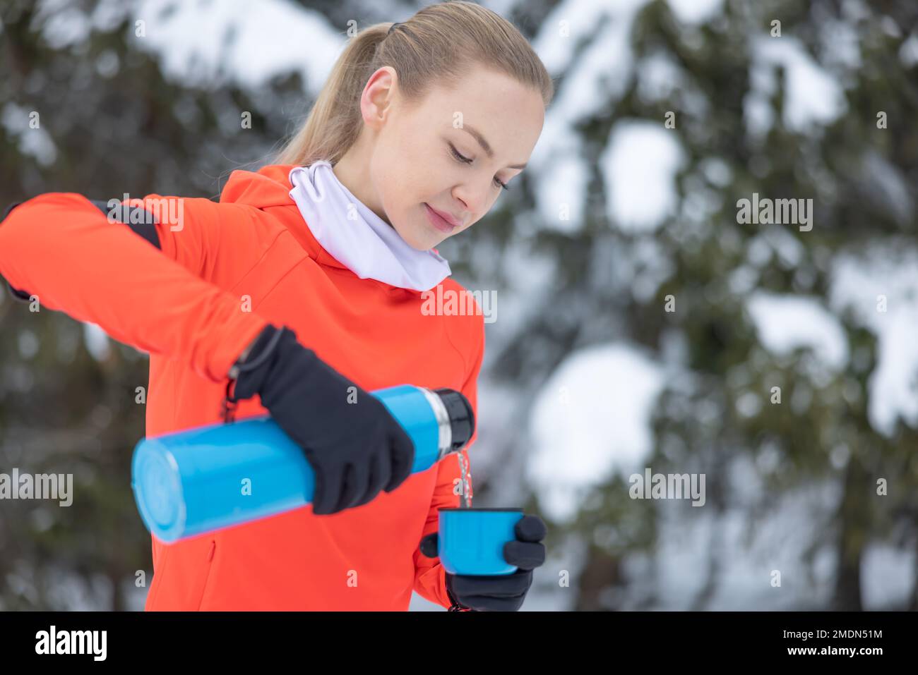 Young fit woman in warm fitness clothes pours hot tea into mug from metal thermos, after workout outdoors on sports ground on winter. Stock Photo