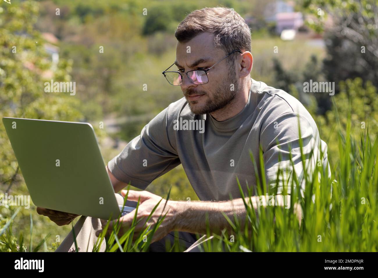 Young adult man with glasses sitting in green grass and working on laptop. Stock Photo