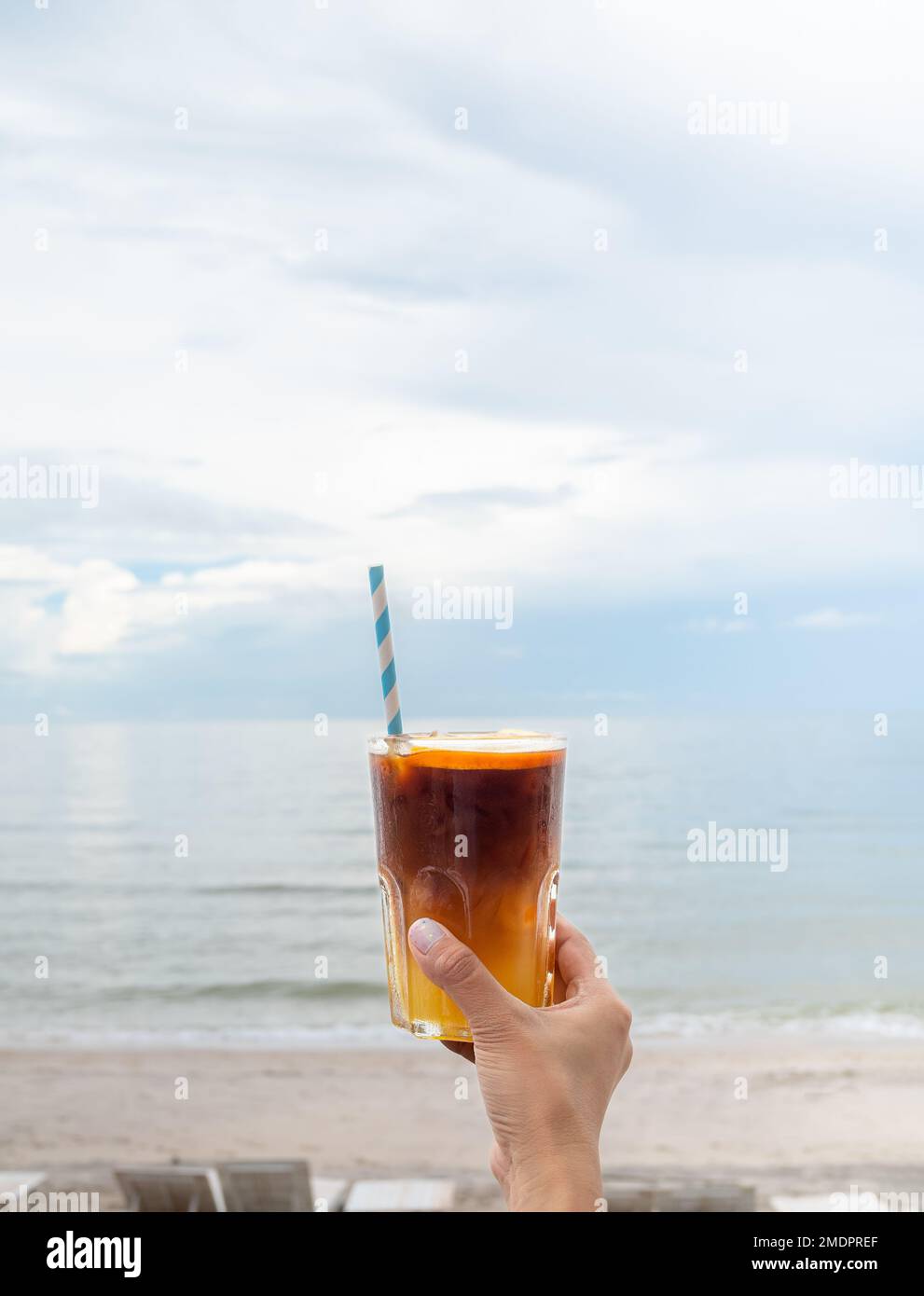 A glass of fresh iced orange americano decorated with sliced orange holding by hand on the beach view, seascape, vertical style. Cold black coffee w Stock Photo