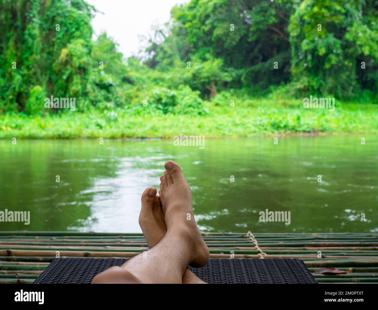 Take a break with relaxing time. The crossed feet of a person resting on a sunbed on a bamboo raft, watching streams in the calm peaceful green forest Stock Photo