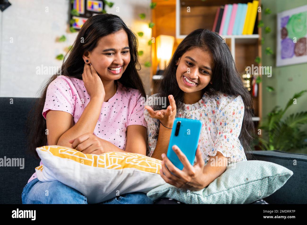 Happy smiling sibling girl sisters on sofa making video call on mobile phone at home - concept of relationship, wireless communication and cyberspace. Stock Photo