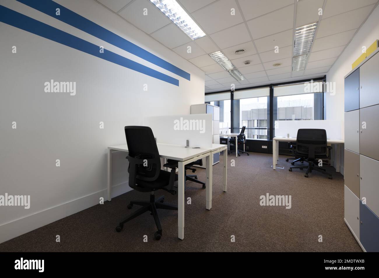 Office with small opposite white tables with partitions, filing cabinet with locks and black swivel chairs Stock Photo