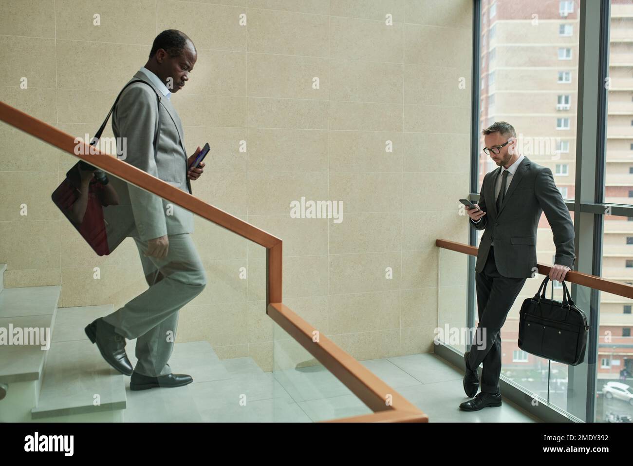 Mature African American businessman walking downstairs while young male entrepreneur using smartphone inside modern business center Stock Photo