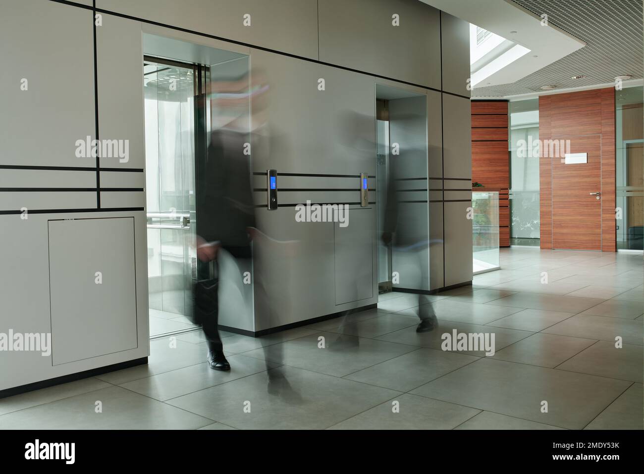 Part of spacious corridor inside modern office center and two blurred businessmen by elevators with glass doors moving along Stock Photo