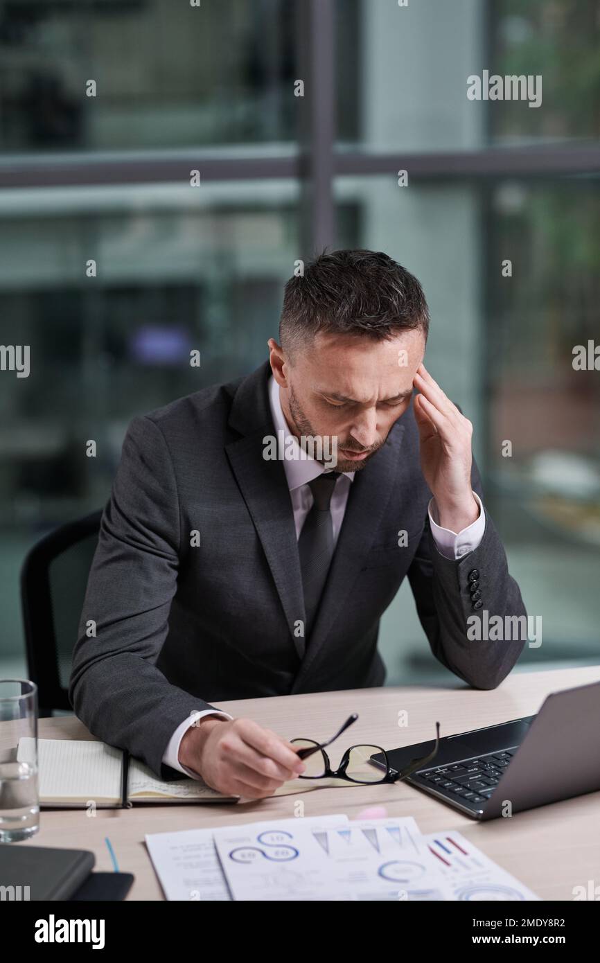 Overworked chief executive office with hand on temple feeling sick while sitting in front of laptop by workplace and analyzing online data Stock Photo