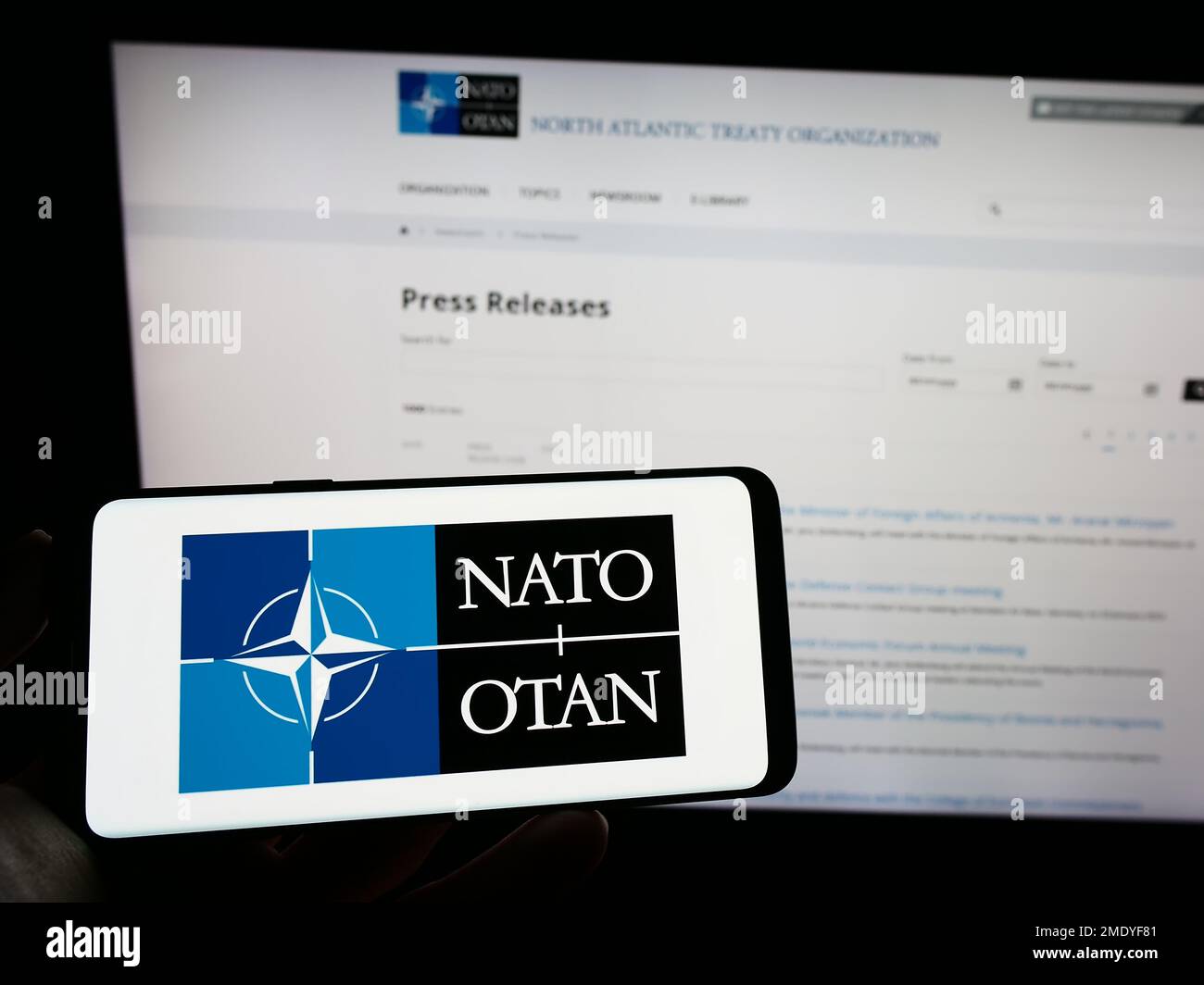 Person holding cellphone with logo of North Atlantic Treaty Organization (NATO) on screen in front of webpage. Focus on phone display. Stock Photo