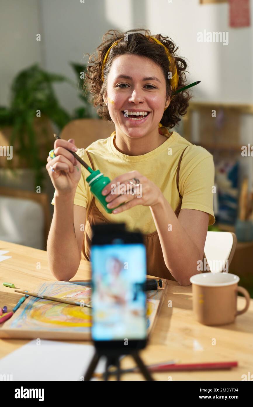 Young cheerful woman painting in front of smartphone while sitting by table with all necessary supplies and giving online masterclass Stock Photo