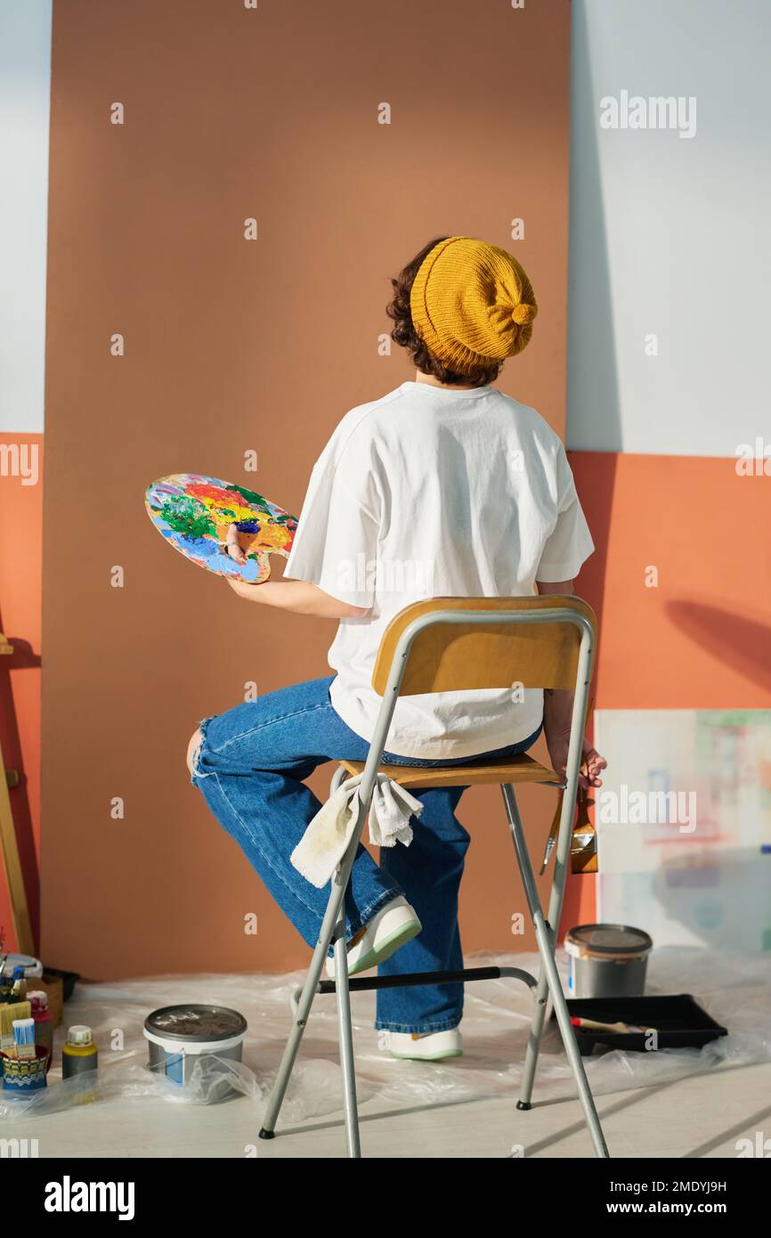 Back view of young creative female painter holding palette with acrylic paints of various colors while working over new masterpiece Stock Photo
