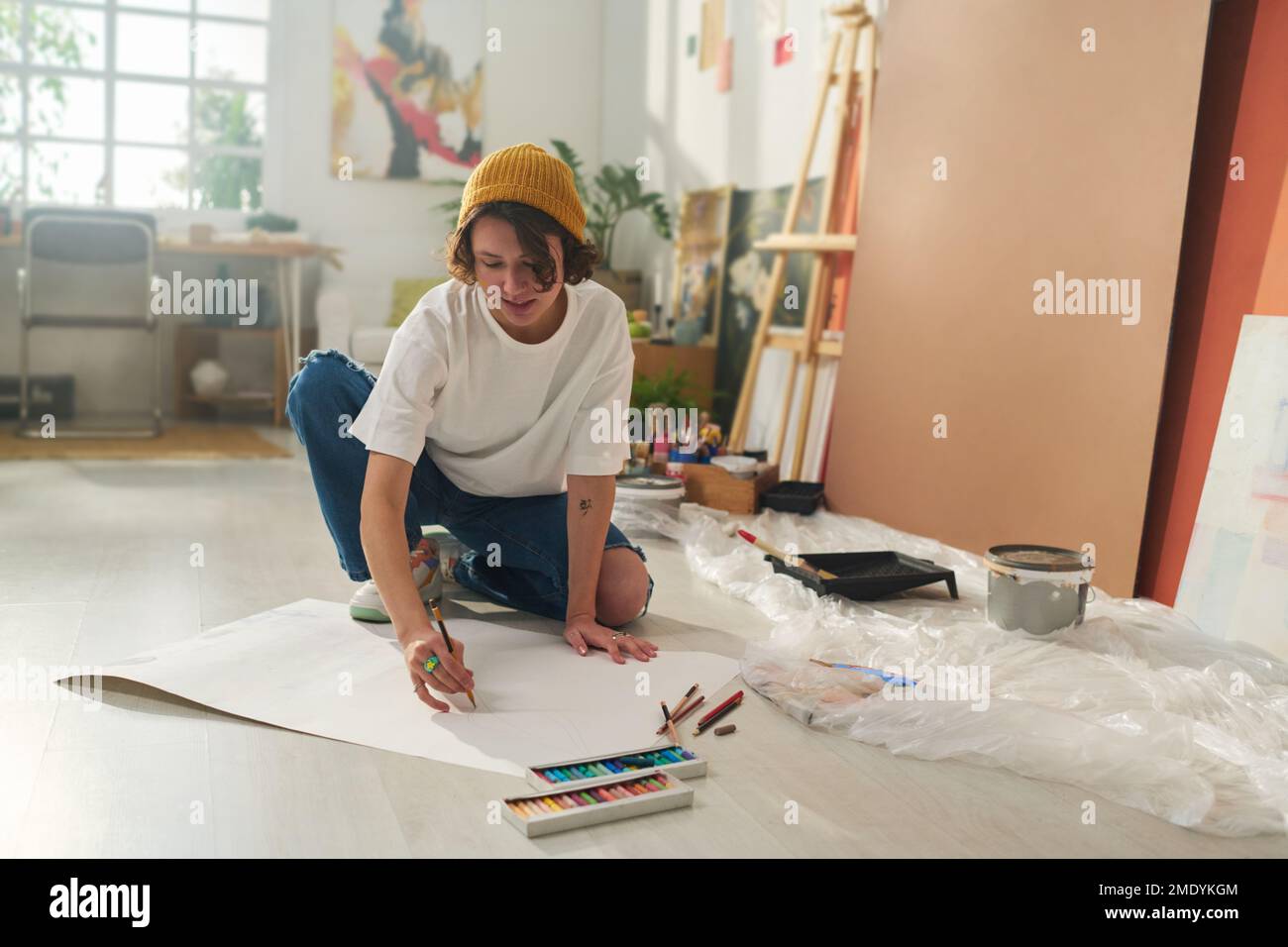 Pretty girl in casualwear sitting on the floor and drawing with crayons while slightly bending over large sheet of white blank paper Stock Photo
