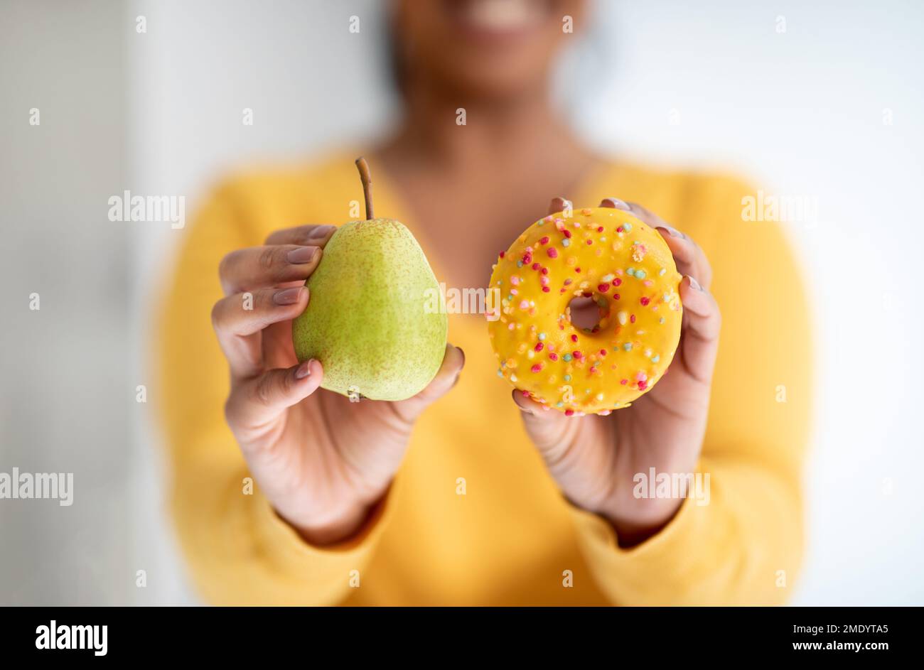 Smiling young african american lady in yellow clothes show pear and donut in minimalist kitchen interior, close up Stock Photo