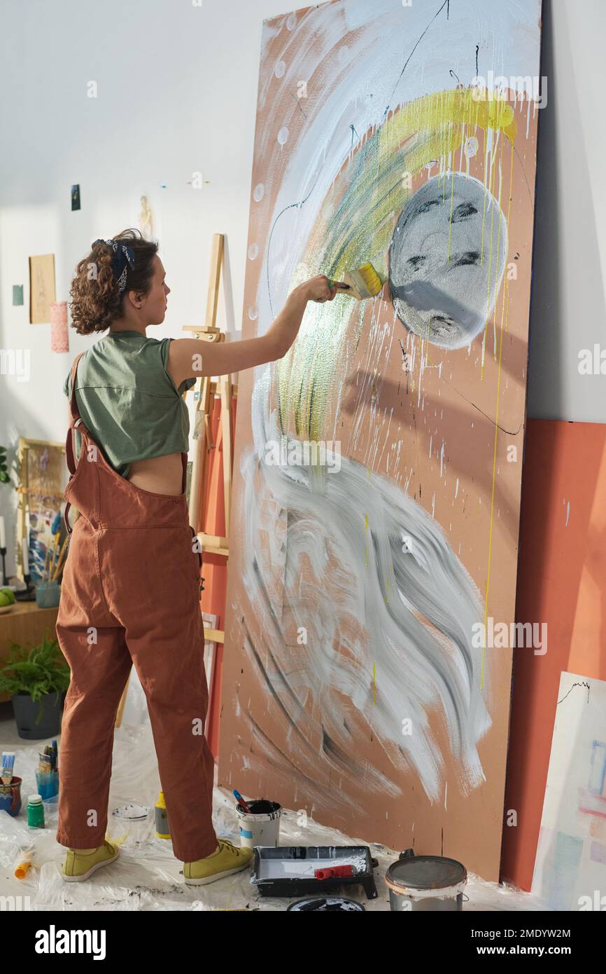 Young craftswoman in casualwear standing in front of panel while painting space theme with moon, starts and galaxy stuff in workshop Stock Photo