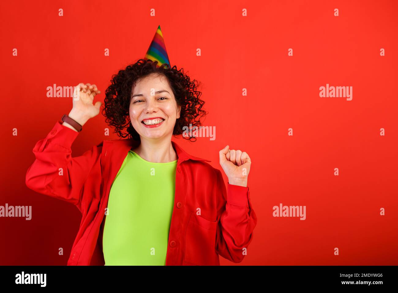 Curly cute girl laughs and jumps in a colorful birthday cap. Studio portrait of white woman on a bright red background. Positive smiling young female. Stock Photo