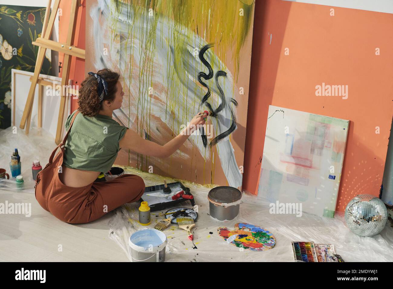 Creative girl in casual attire drawing black outline on grey abstraction on large panel for painting while finiching up with new masterpiece Stock Photo