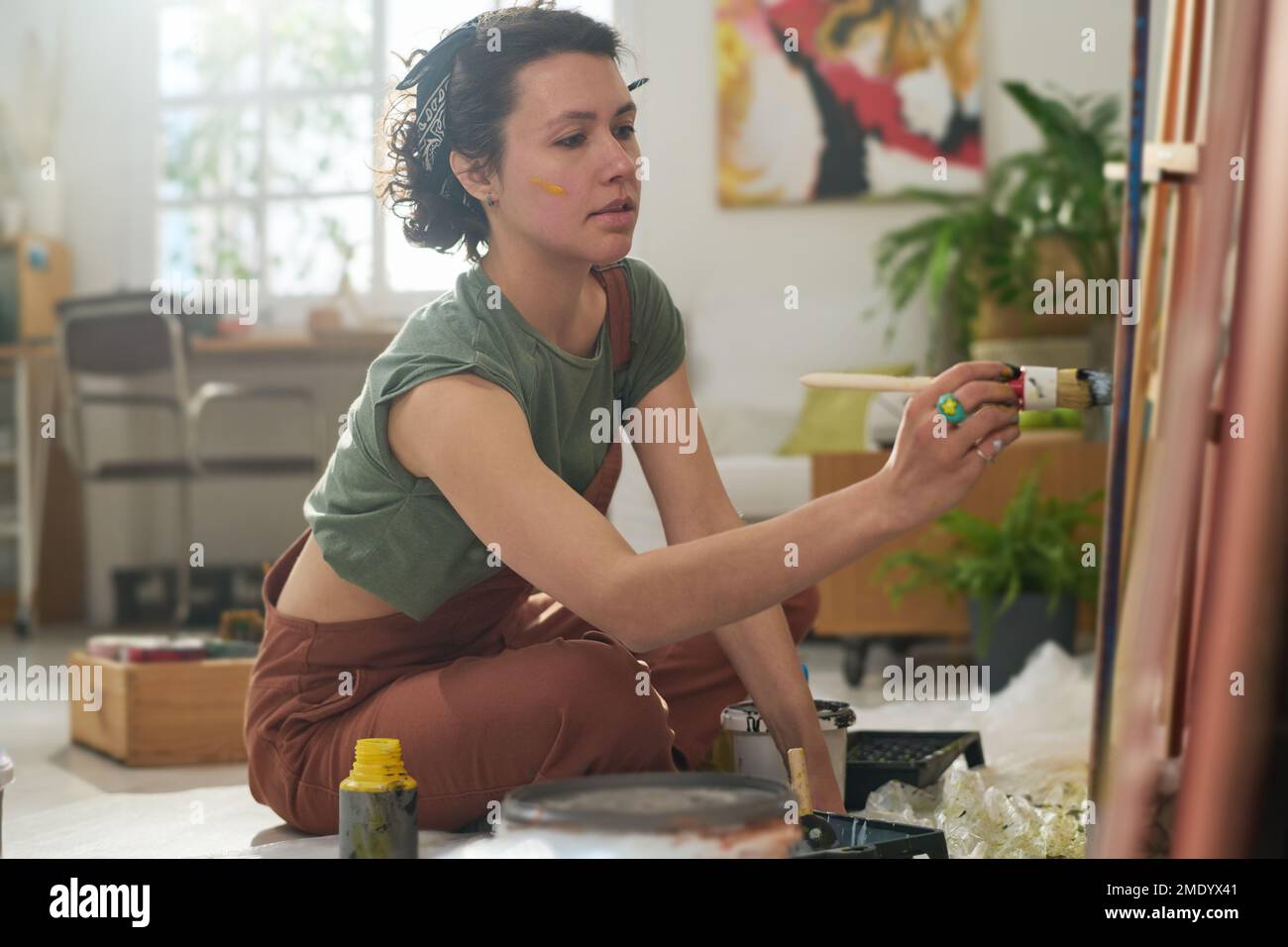 Young serious woman in casual apparel holding paintbrush during process of creating new artwork while sitting on the floor in studio Stock Photo