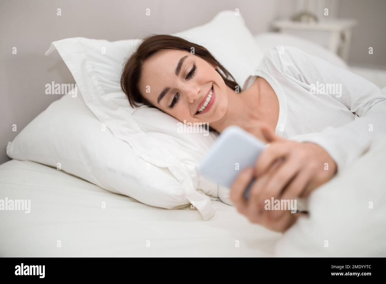 Happy young lady holding smartphone while lying in bed Stock Photo